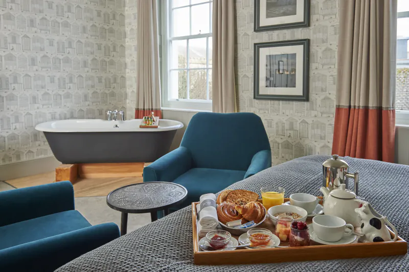 Hotel room with breakfast tray on bed, blue velvet chairs, freestanding bathtub, and patterned wallpaper