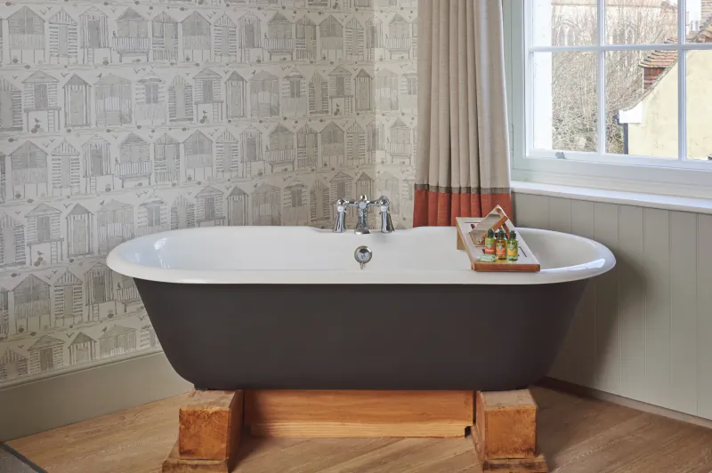 Freestanding black and white bathtub on wooden blocks in a bathroom with beach hut wallpaper and a window