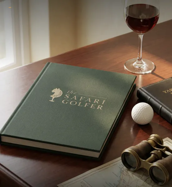 Green hardcover book titled 'The Safari Golfer' with horse logo on dark wood table beside wine glass, golf ball, binoculars, and map