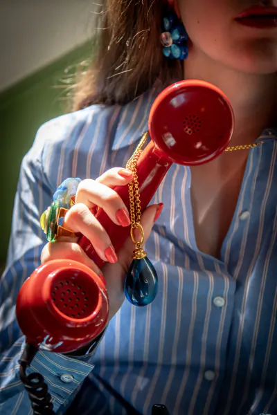 Close-up of a woman in a blue striped shirt holding a red vintage telephone receiver with a gold chain and blue pendant attached
