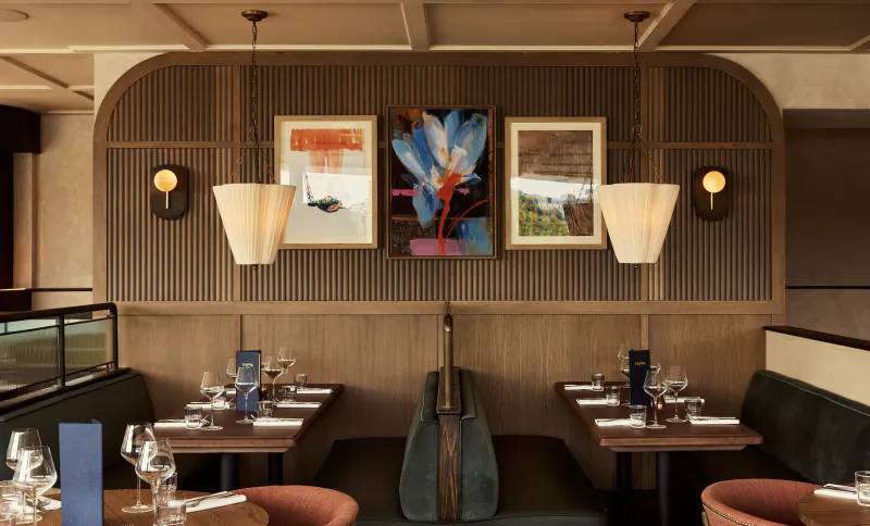 Modern restaurant interior with wooden tables set for dining, three framed artworks on slatted wall, pendant lights, and a large guitar sculpture between booths