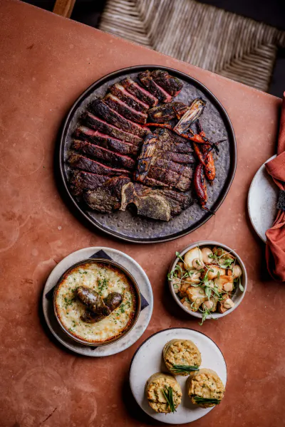 Overhead view of grilled steak platter with sides of mac and cheese, mushrooms, and herb biscuits on a terracotta table