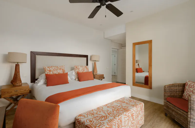Modern hotel bedroom with white bed, orange pillows and runner, wooden headboard, ceiling fan, mirror, and woven armchair