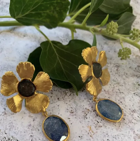Gold flower-shaped earrings with dark and blue gemstones, displayed on a marble surface with green leaves