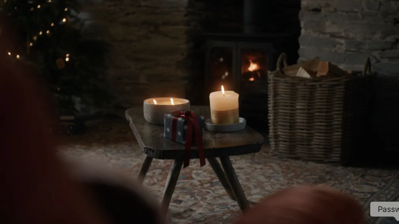 Lit candles and small gift box on wooden table in front of stone fireplace with Christmas tree