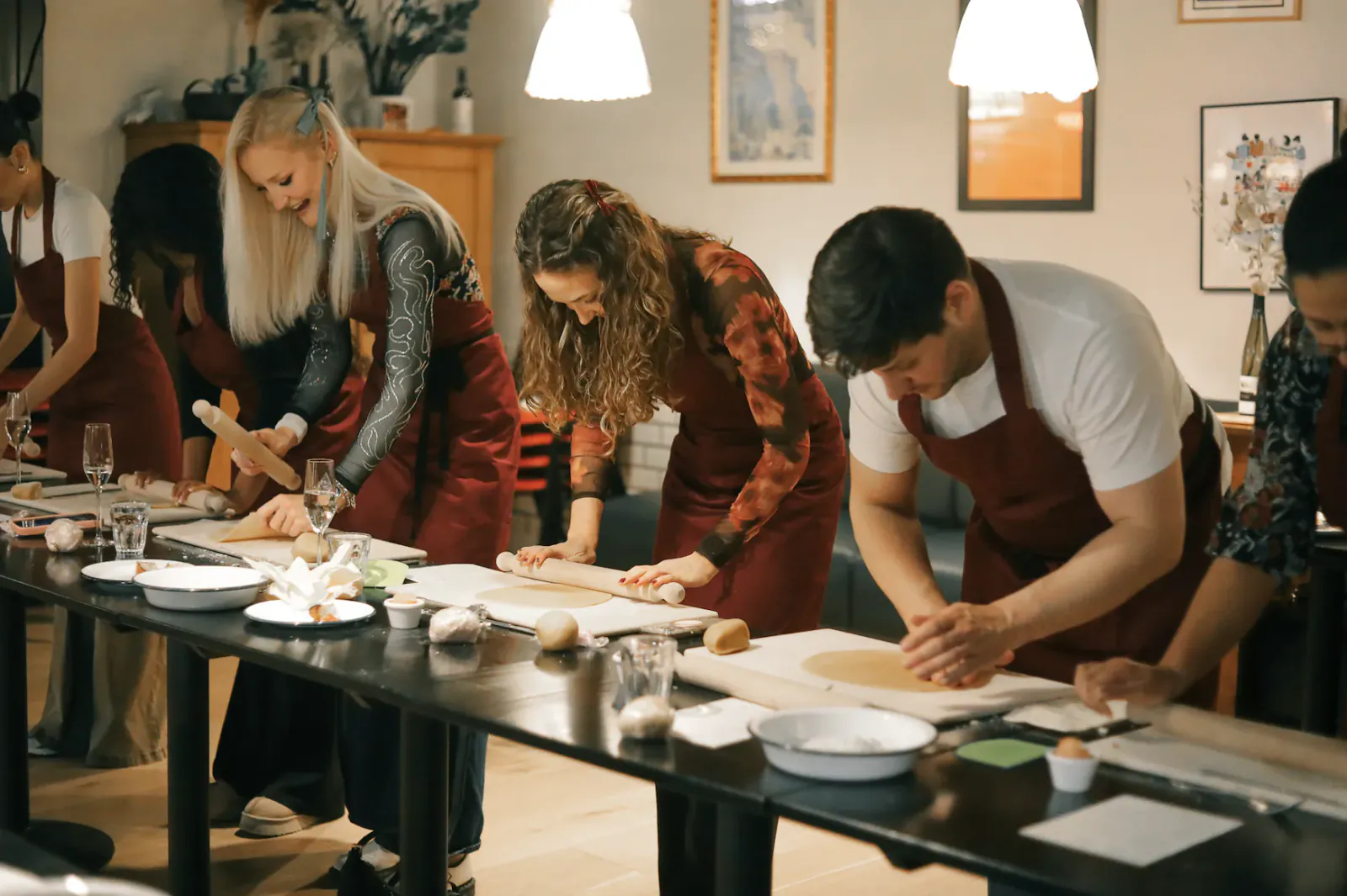 Group of people in red aprons rolling dough at a long table during a pasta-making class