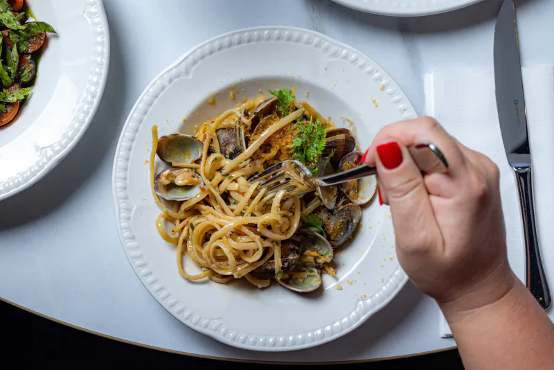 Hand with red nail polish twirling linguine with clams on a white plate using a fork, beside a side salad and knife on a white table