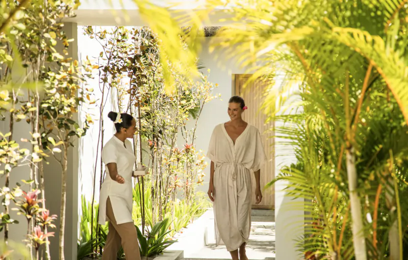Woman in white robe walking through tropical resort hallway with staff member holding tray