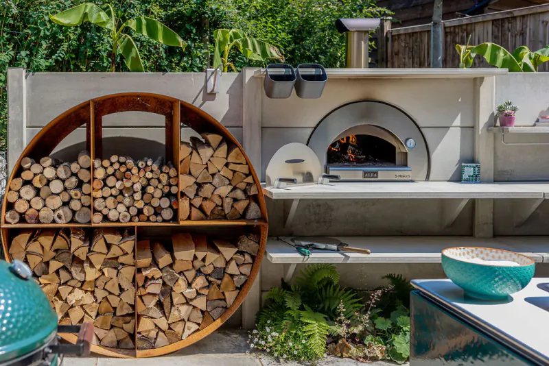 Round wooden rack filled with stacked firewood beside a lit outdoor pizza oven on a patio