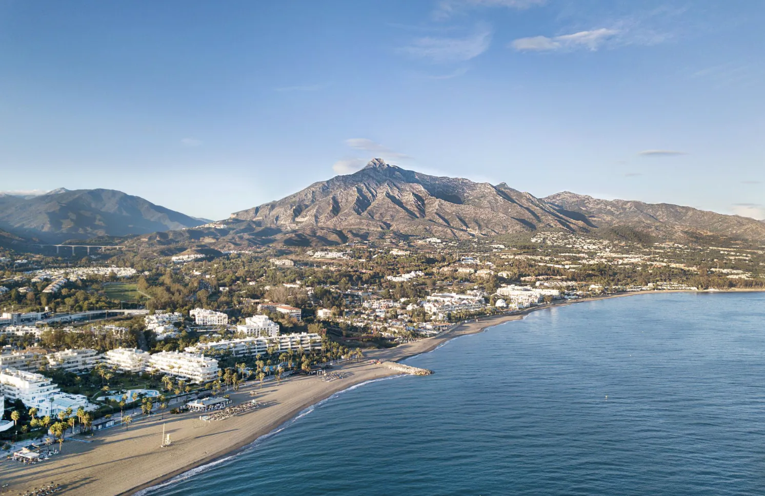Aerial view of Marbella coastline with sandy beach, white buildings, and rocky mountains under clear blue sky