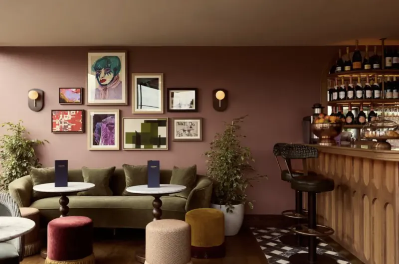 Modern lounge with green velvet sofa, round tables, bar stools, wine bottles on shelves, and gallery wall on dusty rose backdrop