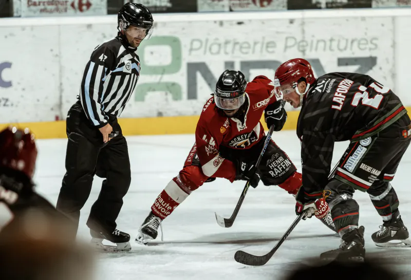 Ice hockey referee watches two players in red and black jerseys battle for puck on rink