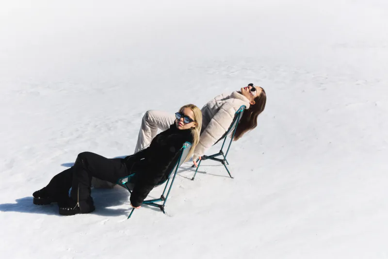 Two women in sunglasses and winter coats relax on folding chairs in a snowy landscape
