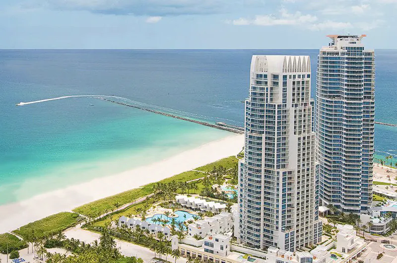 Aerial view of two tall luxury high-rise buildings beside a white sand beach and turquoise ocean with a long pier extending into the water