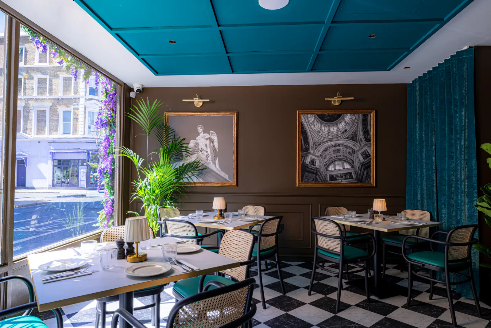 Elegant restaurant interior with teal ceiling, brown walls, checkered floor, and tables set for dining beside large windows with purple flowers