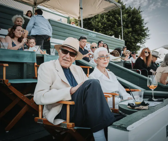 Elderly couple in sunglasses and light jackets seated in director chairs at an outdoor event with spectators in the background
