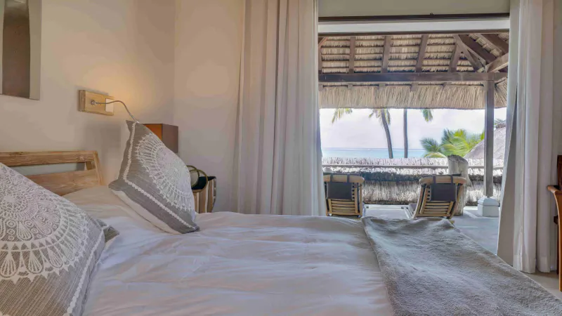 Bright bedroom with white bed, patterned pillows, and open doors overlooking a thatched-roof terrace with ocean view