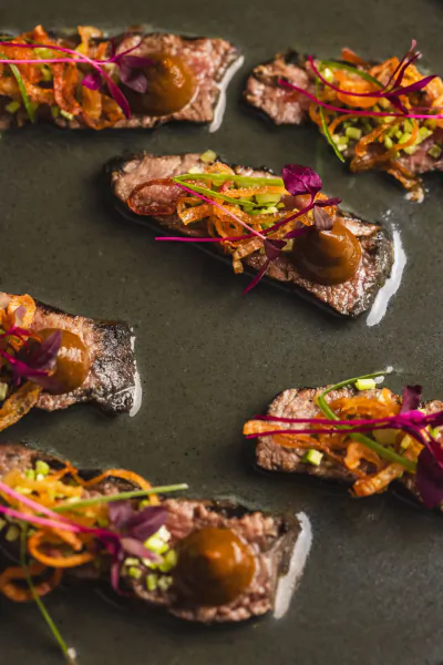 Close-up of gourmet beef canapés on a dark plate, topped with brown sauce, shredded vegetables, microgreens, and edible flowers