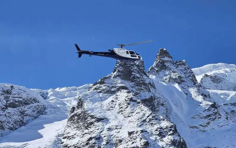 White and black helicopter flying over jagged snow-covered mountain peaks under clear blue sky