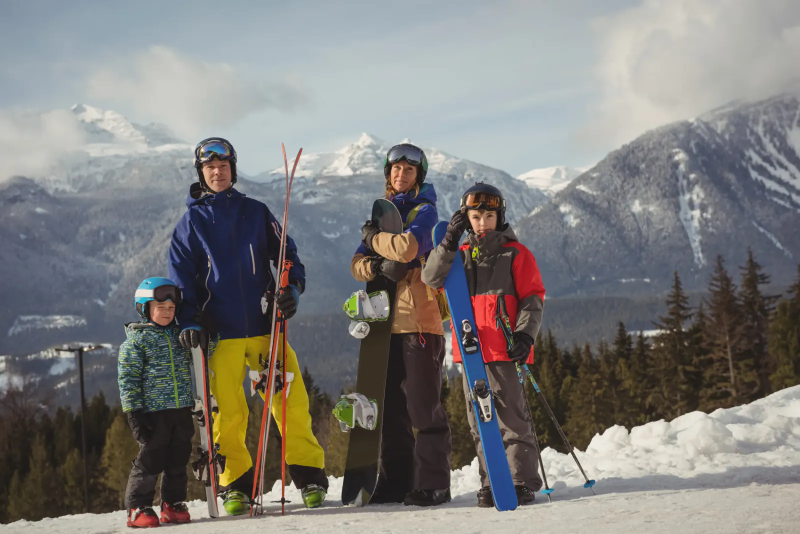 Four skiers, including two adults and two children, posing with skis and snowboards on a snowy mountain slope with peaks in the background