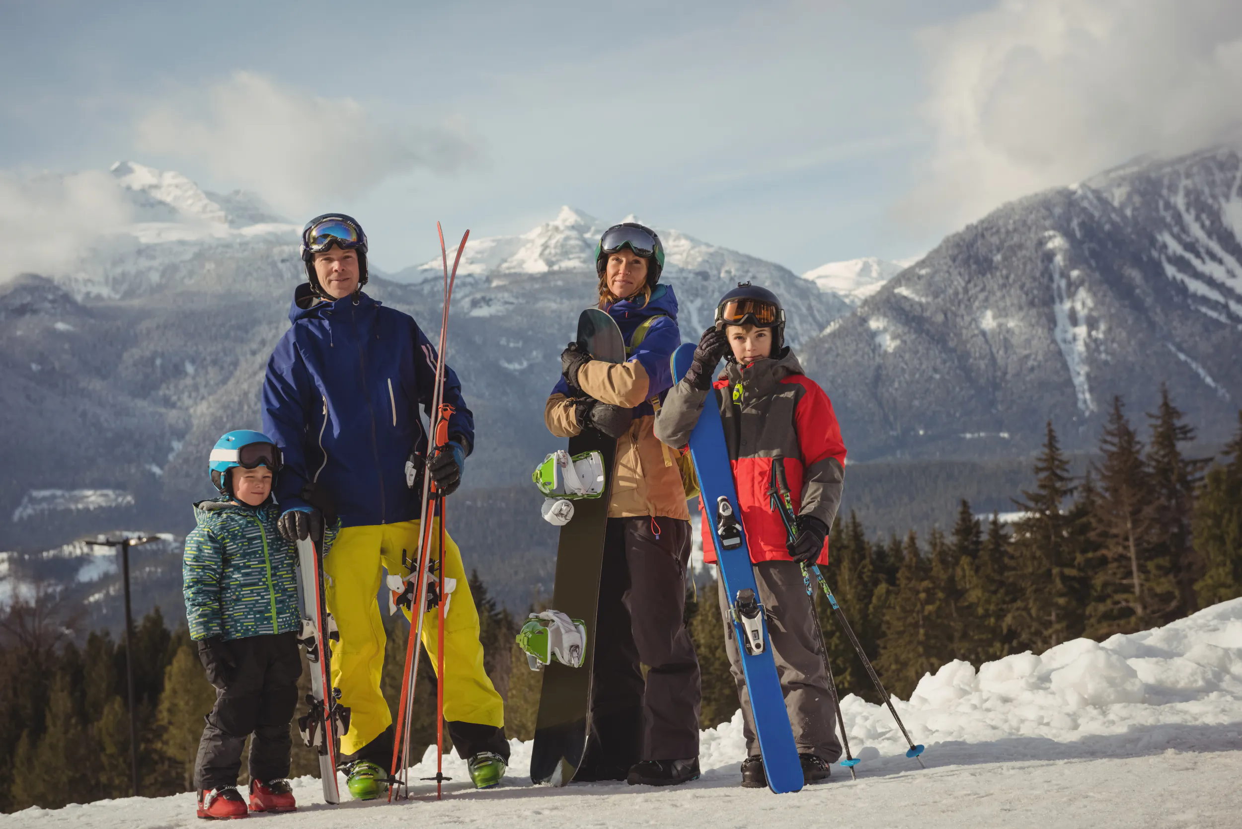 Four skiers, including two adults and two children, posing with skis and snowboards on a snowy mountain slope with peaks in the background