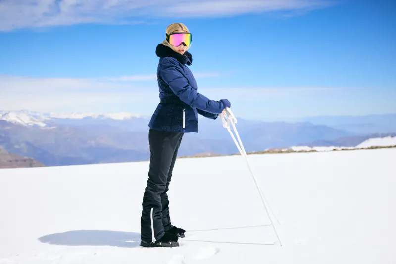 Woman in blue ski jacket and pink goggles holding poles on snowy mountain slope