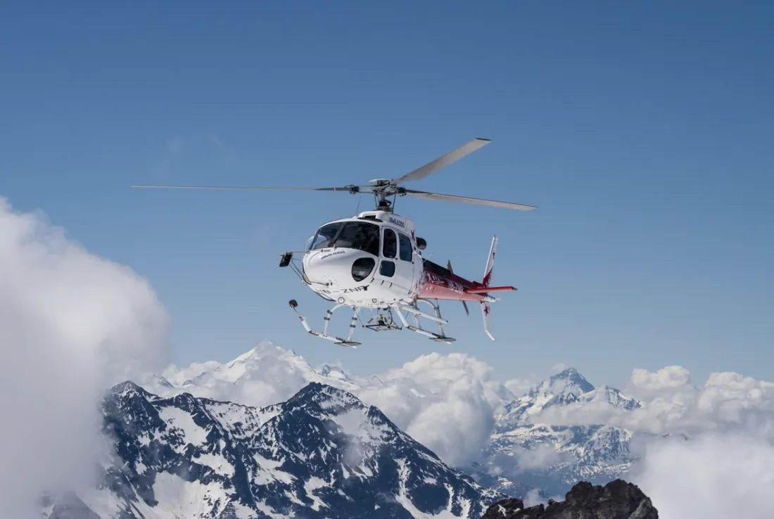 White and red helicopter flying over snow-covered mountain peaks under a clear blue sky