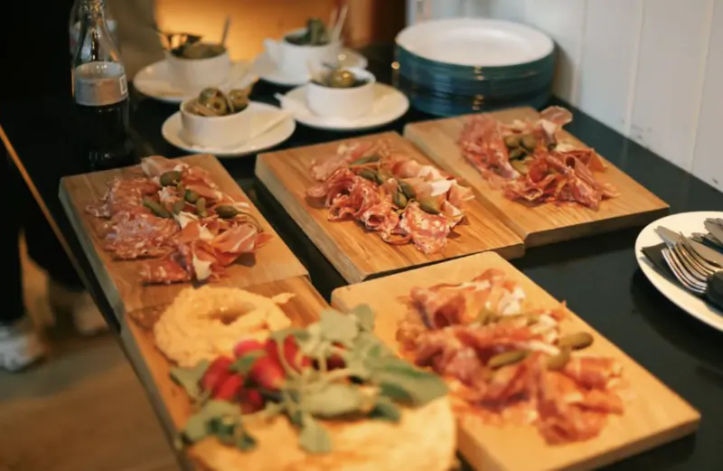 Wooden boards with cured meats, olives, and hummus topped with tomatoes and greens on a restaurant table