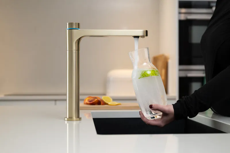 Person filling a glass pitcher with water from a brushed gold kitchen faucet