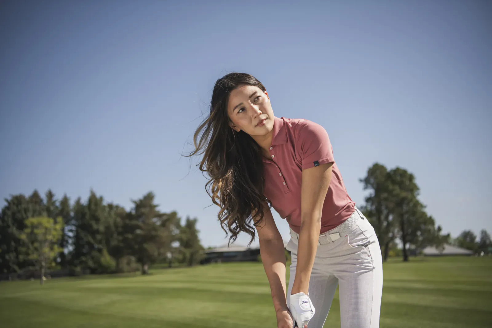 Woman golfer in pink polo shirt and white pants leaning forward on a sunny golf course