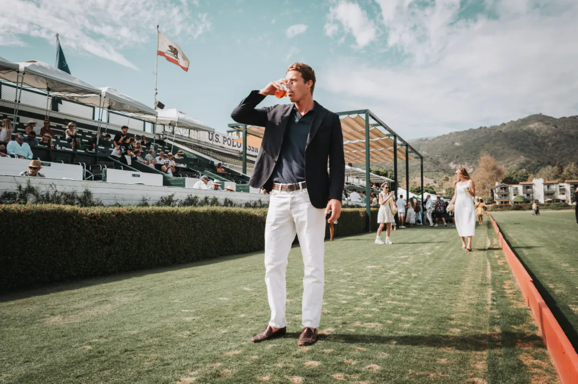 Man in navy blazer and white trousers drinking from a cup on a polo field with spectators and California flag in background