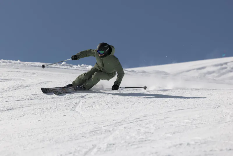 Skier in green KJUS suit carving a turn on snowy slope under clear blue sky