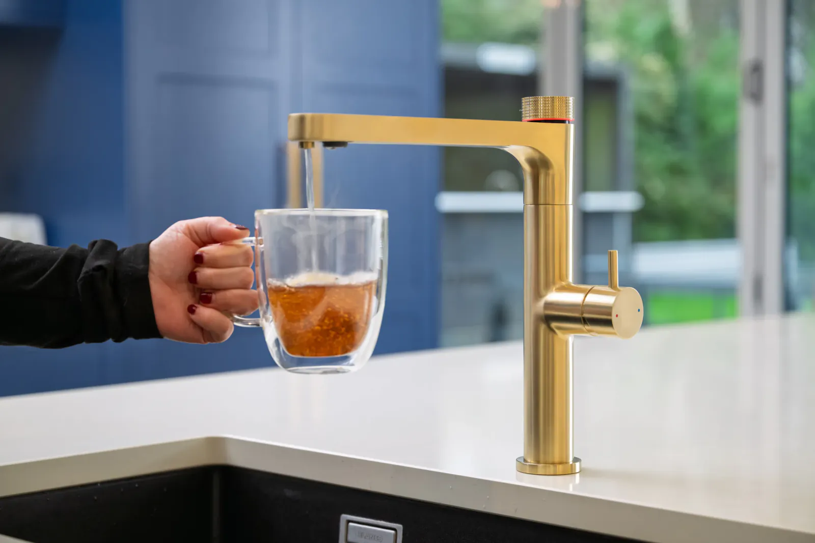 Hand holding double-wall glass mug under gold kitchen faucet filling with amber liquid