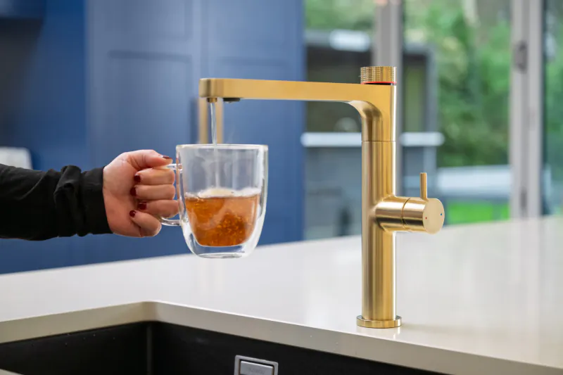 Hand holding double-wall glass mug under gold kitchen faucet filling with amber liquid