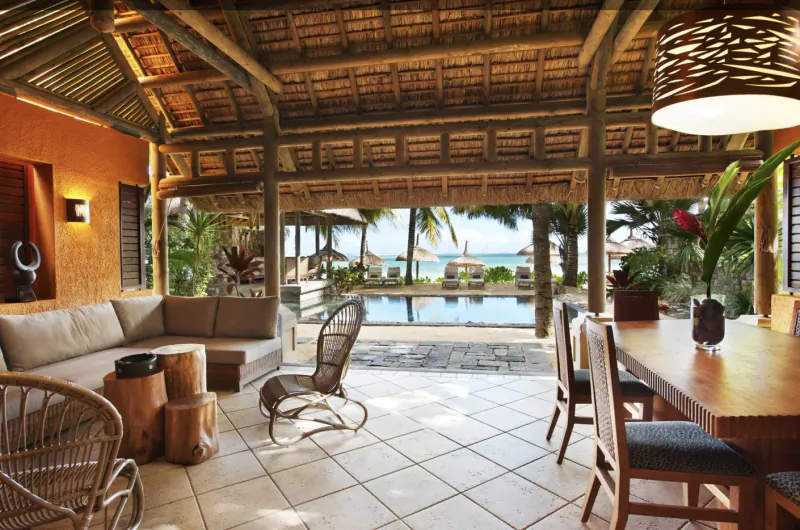 Open-air luxury villa lounge with thatched roof, ocean view, pool, wooden dining table, and rattan seating