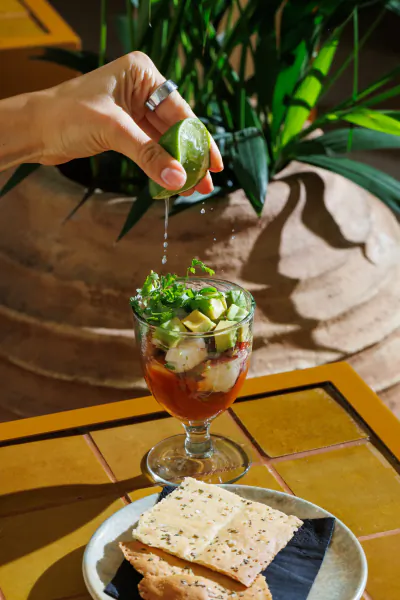 Hand squeezing lime over a layered Mexican shrimp cocktail in a glass, garnished with avocado and herbs, beside crackers on a tiled table