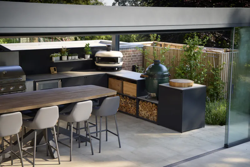 Modern outdoor kitchen with wooden dining table, four grey bar stools, built-in grills, green ceramic smoker, and firewood storage