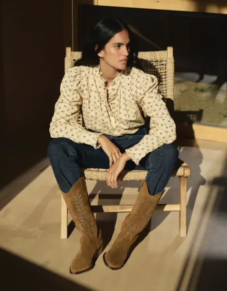 Woman in floral blouse and cowboy boots sitting on woven chair indoors