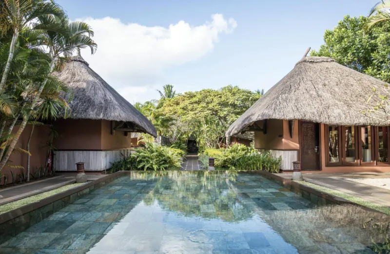 Luxury resort villas with thatched roofs beside a tranquil pool surrounded by tropical palms and lush greenery under a bright sky