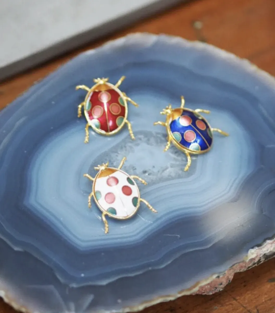 Three colorful enamel ladybug brooches with gold trim displayed on a blue agate slice