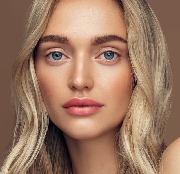 Close-up portrait of a woman with long wavy blonde hair, blue eyes, and natural makeup against a neutral brown background