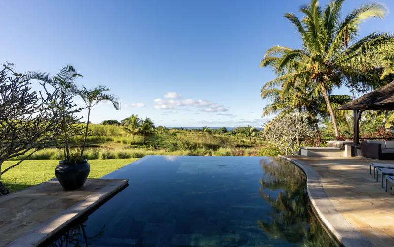 Infinity pool overlooking lush green fields and ocean at Heritage The Villas Mauritius under clear blue sky