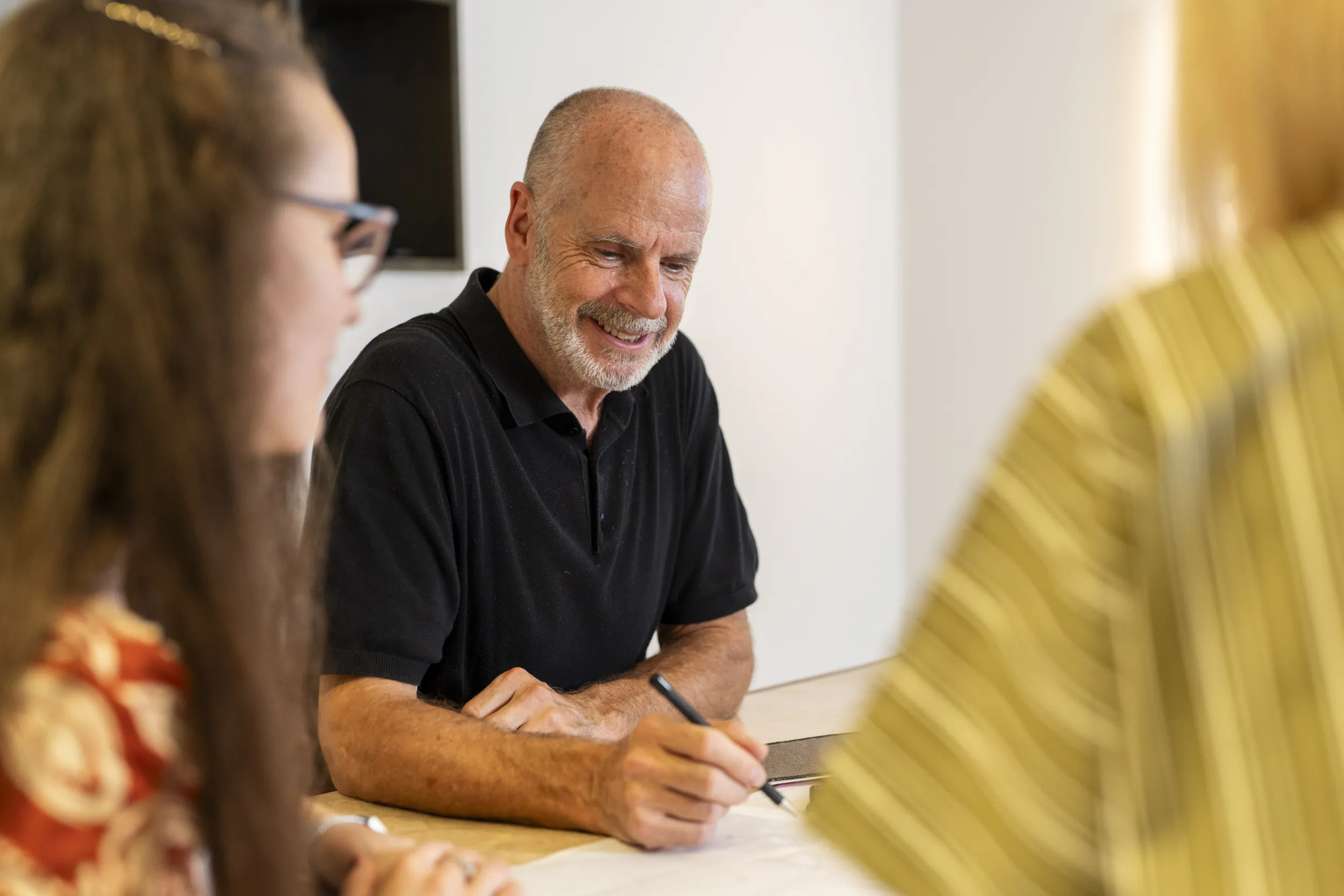 Smiling man in black polo shirt writing at table during interview with two women