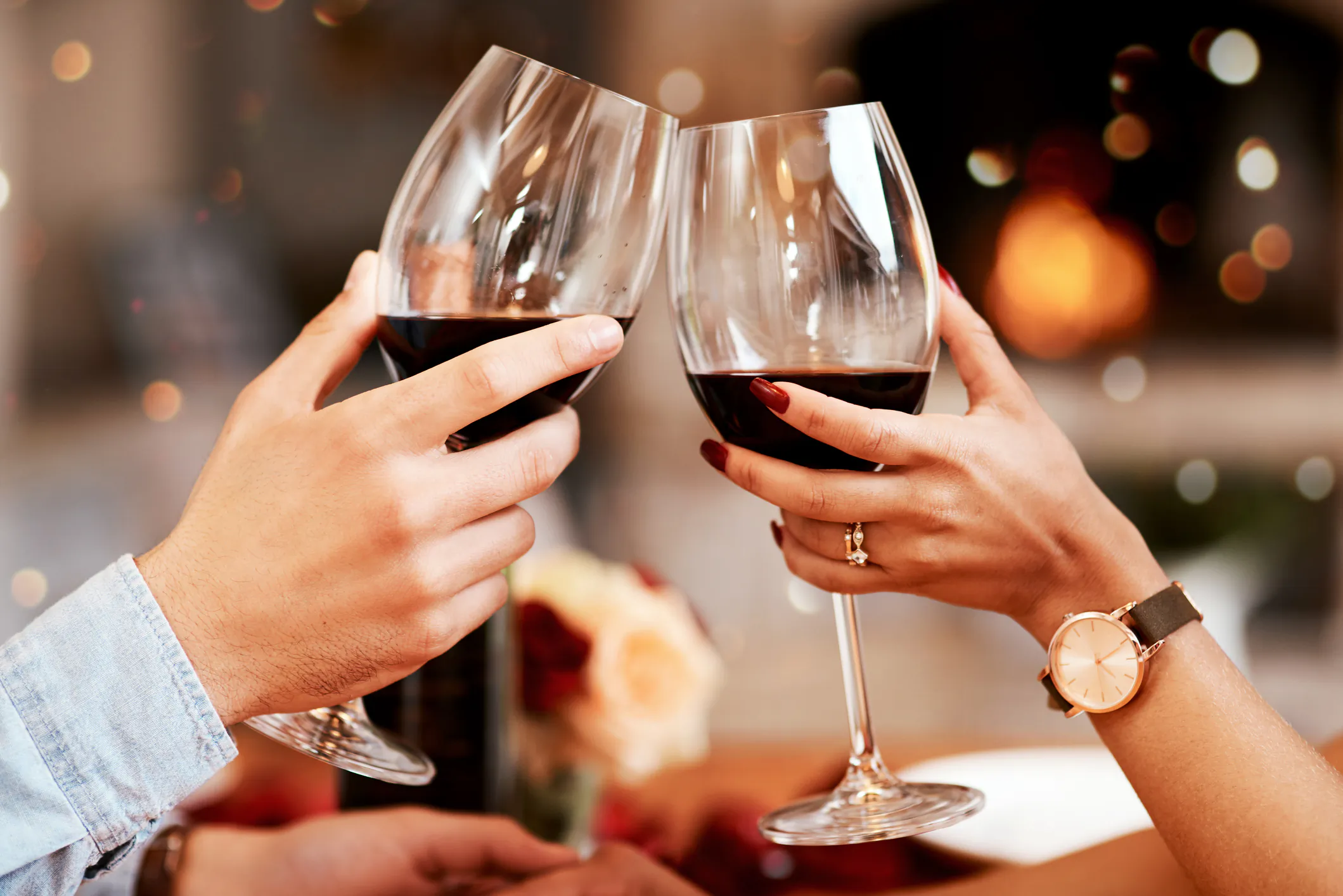Two hands clinking red wine glasses in a romantic toast, one wearing a watch and ring