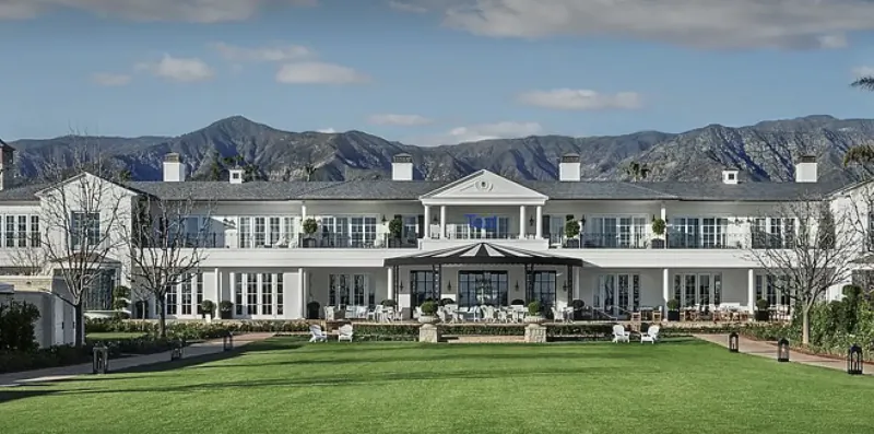 Large white mansion with dark roof overlooking green lawn and mountains