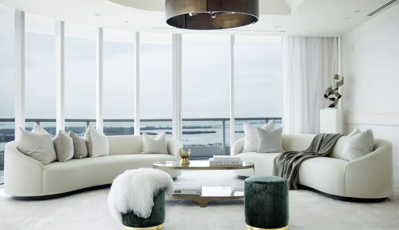 Modern luxury living room with curved white sectional sofa, green velvet ottomans, and floor-to-ceiling windows overlooking water