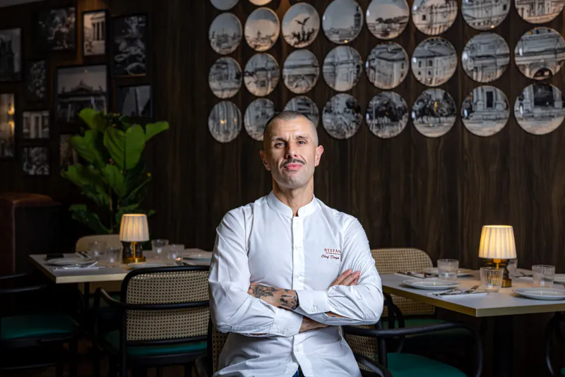 Chef Diego, a man in a white chef's jacket with arms crossed, seated in a stylish restaurant with decorative plates on the wall