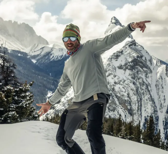 Man in green beanie and sunglasses pointing excitedly on snowy mountain peak