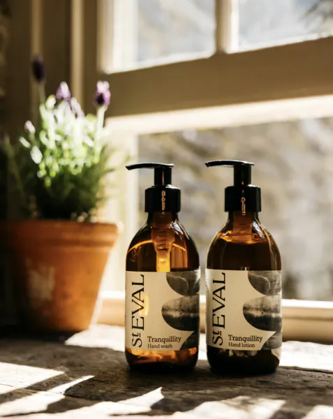 Two amber glass bottles of St Eval Tranquillity hand wash and hand lotion on a wooden windowsill with sunlight and a potted plant