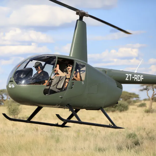 Green helicopter ZT-RDF flying low over savanna with two pilots in cockpit, one waving